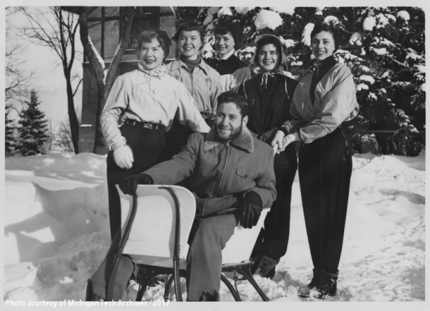 The 1955 Carnival Queen candidates pose outside with a young man in a sled.